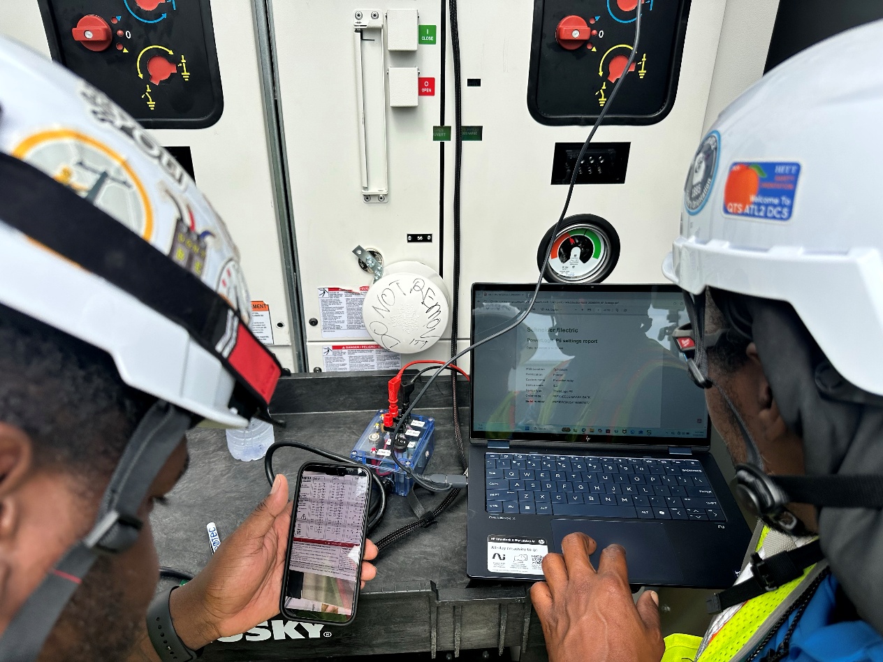 Two developing technicians download protective relay settings together in front of electrical equipment.